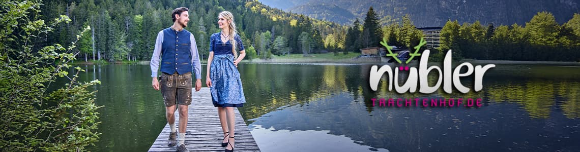 Nübler Trachtenhof – Mann in Lederhose und Frau im blauen Dirndl gehen auf einem Steg am Seeufer, umgeben von Wald und Bergen