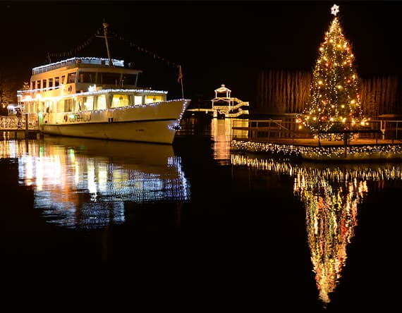 Beleuchtetes Ausflugsschiff und festlich geschmückter Weihnachtsbaum spiegeln sich im nächtlichen Wasser.