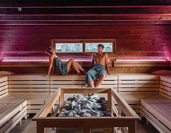 Zwei Personen in Handtüchern sitzen in moderner Sauna mit Holzbänken und Steinen, Fenster mit Blick auf verschneite Landschaft
