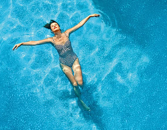 Frau im gestreiften Badeanzug treibt mit ausgebreiteten Armen auf dem Rücken im klaren blauen Pool