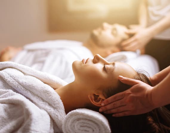 Paar in weißen Bademänteln liegt auf Massageliegen und erhält gleichzeitig eine entspannende Kopfmassage