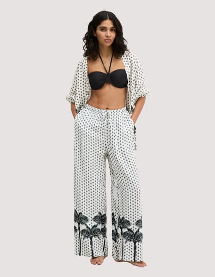 Frau in schwarzem Bikini-Top mit offener gemusterter Bluse und weitem Strandhose mit Palmenprint am Saum