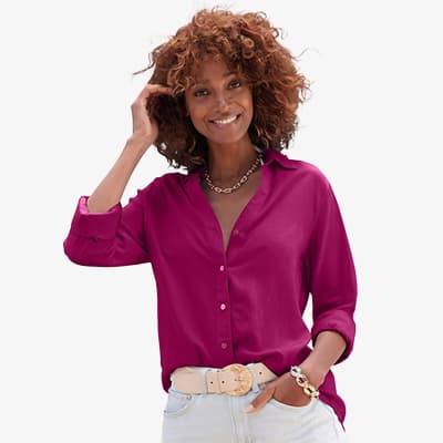 Frau in pinker Langarmbluse und heller Jeans lächelt in die Kamera und hat eine Hand in der Hosentasche
