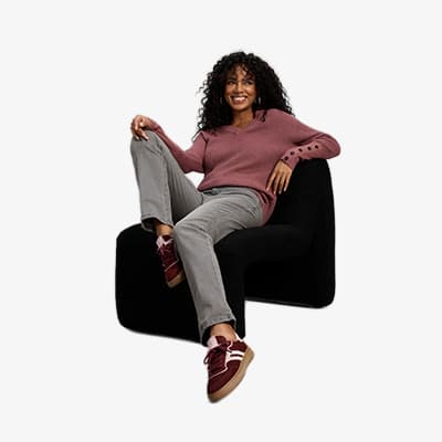 Frau in rosa Pullover und grauer Hose sitzt entspannt auf schwarzem Sessel und zeigt bordeauxrote Sneaker