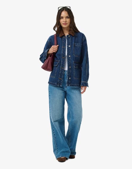 Frau in weiter Jeans und langer Jeansjacke steht frontal und trägt eine bordeauxrote Schultertasche