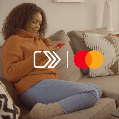Frau mit lockigem Haar in braunem Pullover sitzt auf grauem Sofa und benutzt Smartphone, Mastercard-Logo vorne