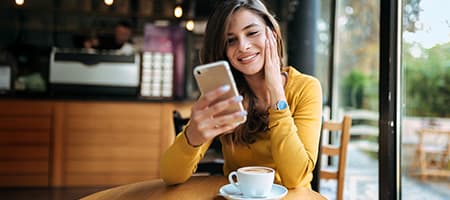 Frau mit gelbem Pullover sitzt in einem Café und schaut auf ihr Smartphone, daneben eine Tasse Cappuccino