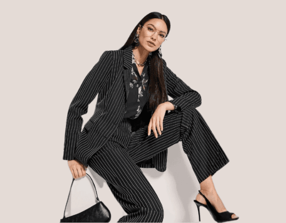 Frau in schwarzem Nadelstreifenanzug und Bluse, sitzt mit Handtasche auf einem Hocker.