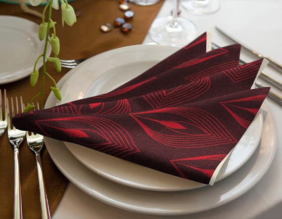 Rote Stoffserviette mit schwarzem Muster, kunstvoll gefaltet auf einem weißen Teller, neben Besteck und Glas auf gedecktem Tisch