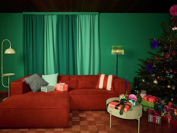 Wohnzimmer mit roten Sofa, grüner Wand und Vorhängen, Weihnachtstannenbaum, Geschenke auf Tisch und unter Baum, Stehlampe links