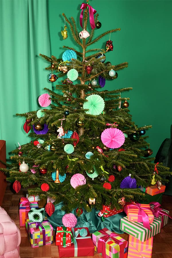 Bunt geschmückter Weihnachtsbaum mit Lametta, Kugeln und Papierrosetten, darunter zahlreiche farbenfrohe Geschenke mit Schleifen