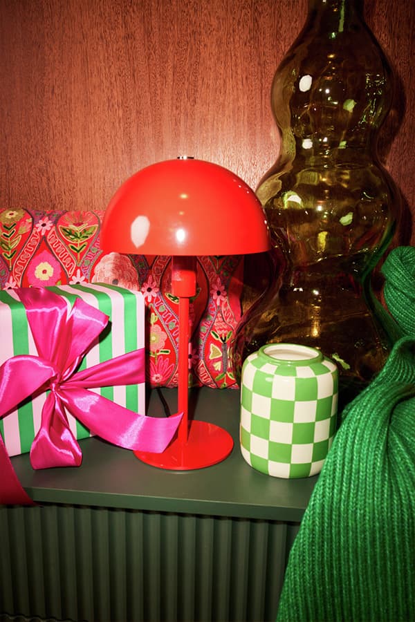 Rote Tischlampe, grün-weißes Schachbrett-Mustergefäß, Geschenk mit grün-weißer Verpackung und rosa Schleife auf grünem Tisch