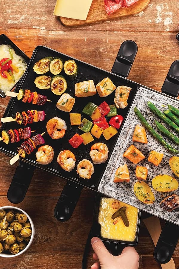 Verschiedene Grillgerichte mit Gemüse, Meeresfrüchten und Käse auf einem Tisch, darunter Oliven und Käsefondue in einer Pfanne