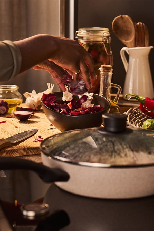 Hand gibt geschnittene rote Zwiebeln in eine schwarze Schüssel, umgeben von Küchenutensilien und frischen Zutaten auf einem Holztisch.