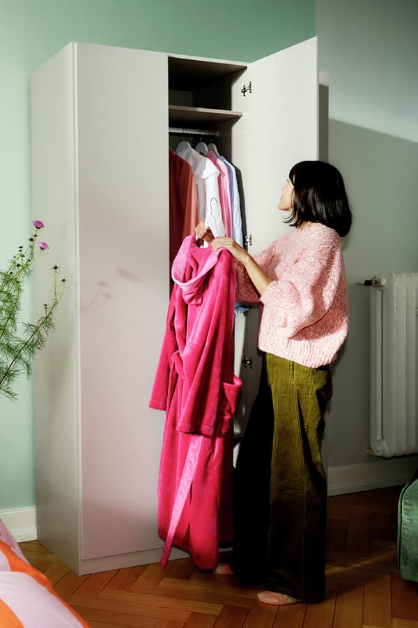Frau in rosa Pullover hängt pinken Mantel in weißen Kleiderschrank. Kleidung hängt auf Bügeln im Schrank.