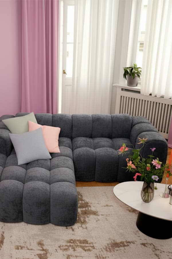 Dunkelgraues Sofa mit bunten Kissen, rundem Tisch und Blumen in einem hellen Wohnzimmer mit rosa Vorhängen.