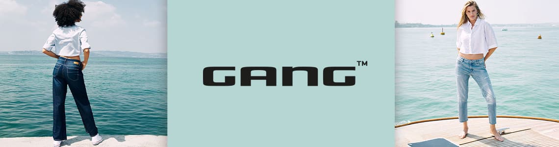 GANG – Zwei Frauen in Jeans stehen am Wasser, eine von hinten, die andere von vorne, beide mit weißem Hemd.