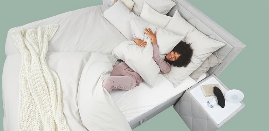 Frau in lila Schlafanzug liegt gemütlich im Bett und umarmt ein Kissen. Auf dem Nachttisch liegen eine Schlafmaske und ein Glas Wasser.