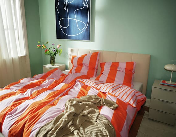 Bett mit orange-weiß gestreifter Bettwäsche, Blumenvase auf Nachttisch, abstraktes Bild an mintgrüner Wand.