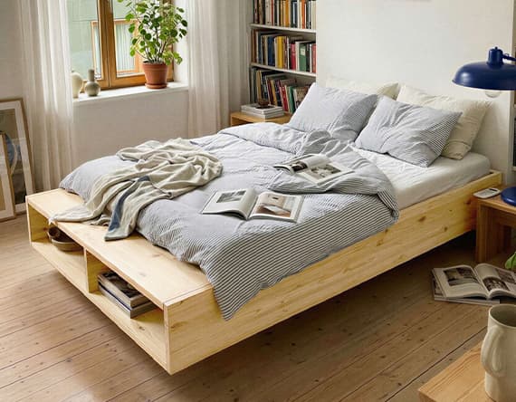 Bett mit grauer Bettwäsche in hellem Zimmer, Bücher und Pflanze auf Fensterbank, Bücherregal im Hintergrund