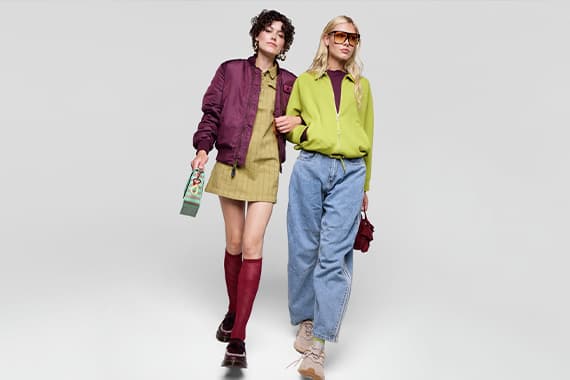 Zwei Frauen gehen Arm in Arm, tragen farbige Freizeitoutfits mit Jacken, Kleid und weiter Jeans vor hellem Hintergrund