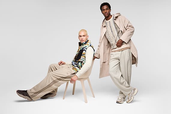 Zwei Männer in hellen Outfits, einer sitzt lässig auf Stuhl, der andere steht daneben im langen Mantel
