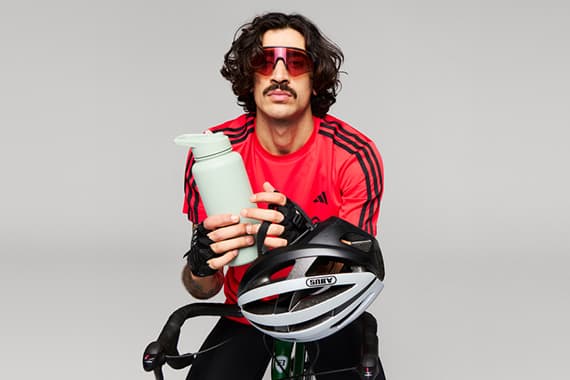 Mann im roten Radtrikot auf Heimtrainer hält Trinkflasche, Fahrradhelm liegt vor ihm am Lenker