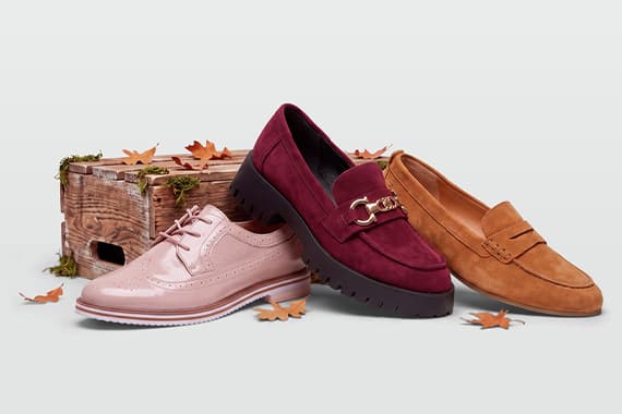 Drei Paar Halbschuhe in Rosa, Bordeaux und Camel vor Holzkiste, umgeben von herbstlichen Blättern
