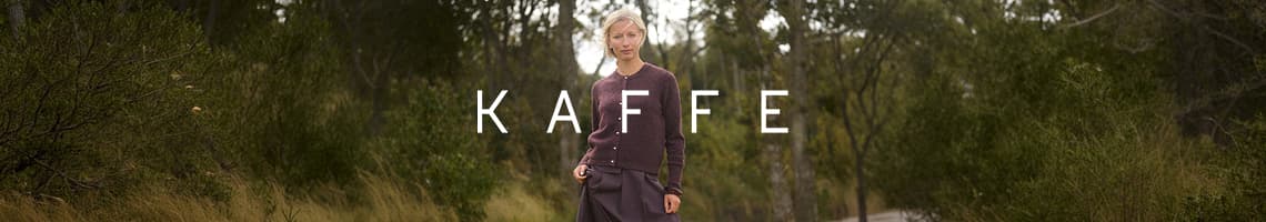 KAFFE – Frau im lila Cardigan steht auf Waldweg und blickt frontal in die Kamera