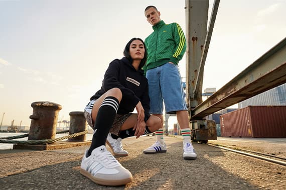 Zwei Personen in sportlichen Outfits posieren am Hafen, Fokus auf weißen Sneakern und gestreiften Socken