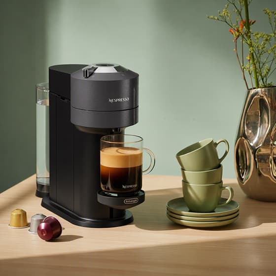 Nespresso Kapselmaschine brüht Kaffee in Glasbecher, daneben Kaffeekapseln und gestapelte Tassen auf Holztisch