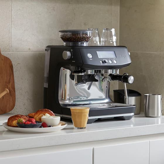 Espressomaschine auf Küchenarbeitsplatte, davor Glas Latte und Teller mit Croissants, Beeren und Joghurt