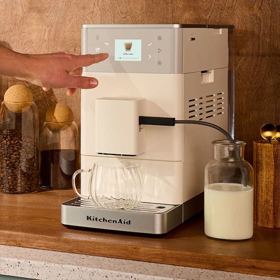 Hand wählt am Display einer KitchenAid Kaffeemaschine „Caffè Latte“, Glas und Milchflasche stehen bereit