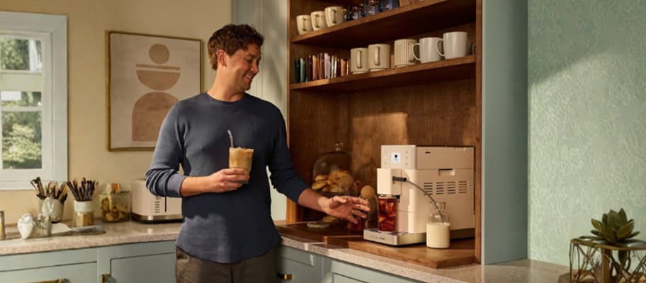 Mann in Küche bereitet mit Kaffeevollautomat ein Getränk zu und hält ein Glas Eiskaffee in der Hand
