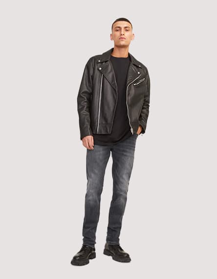 Mann steht frontal in schwarzer Jacke, dunklem Shirt, grauer Jeans und schwarzen Schuhen vor hellem Hintergrund