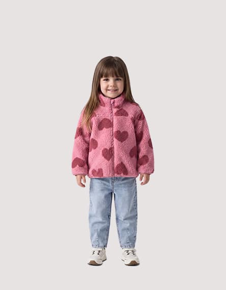 Kind steht frontal zur Kamera in pinker Jacke mit Herzmuster, hellblauer Jeans und Turnschuhen