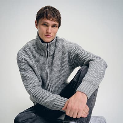 Junger Mann sitzt entspannt auf dem Boden und trägt einen grauen Strickpullover mit hohem Kragen und Reißverschluss