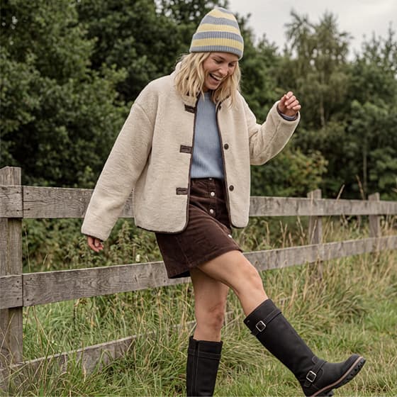 Frau im Herbstoutfit mit Mütze, kurzer Knopfleiste-Rock und Stiefeln läuft lachend auf einer Wiese an einem Holzzaun entlang