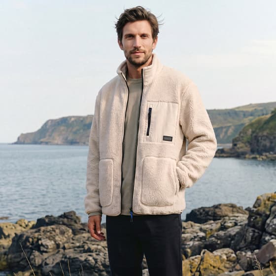 Mann in heller Jacke steht mit Händen in den Taschen auf Felsen vor einer Meeresküste