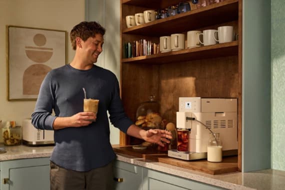 Mann in Küche bereitet mit Kaffeemaschine ein Getränk zu und hält ein Glas Eiskaffee in der Hand