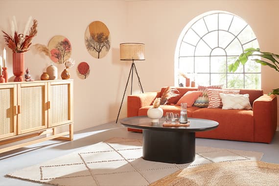 Helles Wohnzimmer mit orangefarbenem Sofa, rundem Fenster, Dekokissen, schwarzem Couchtisch und Sideboard mit Vasen