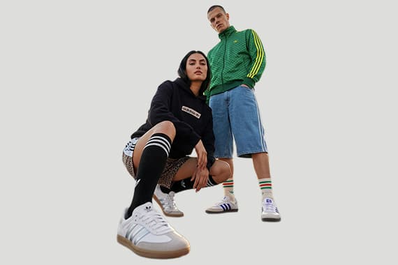 Zwei Personen in sportlicher Streetwear, eine hockt im Vordergrund, die andere steht dahinter in Shorts und Jacke