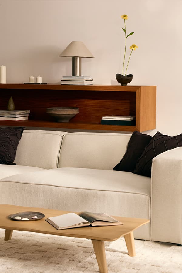 Helles Sofa mit dunklen Kissen vor Holzregal, davor niedriger Tisch mit offenem Buch und Schale