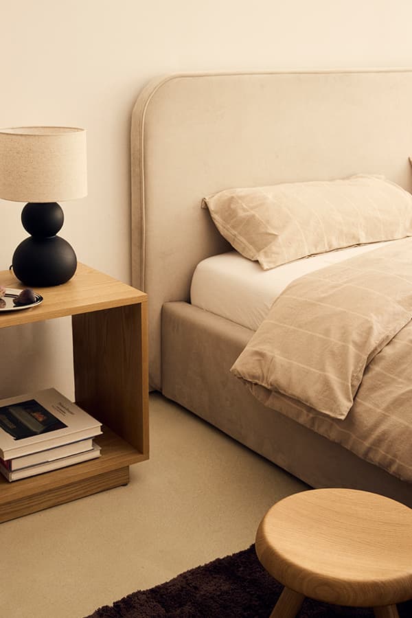 Bett mit beiger Bettwäsche, Nachttisch mit Lampe und Büchern, davor kleiner Hocker auf Teppich