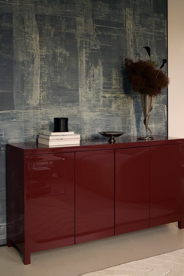 Dunkelrotes Sideboard vor blaugrauer Wand, darauf Bücherstapel, schwarze Dose, Schale und Vase mit Gräsern