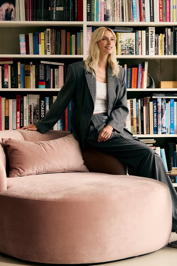 Frau im Nadelstreifenanzug sitzt auf rundem rosa Sofa vor einem vollen Bücherregal