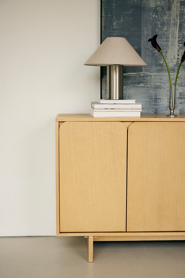 Helles Sideboard mit gerundeten Türen, darauf Tischlampe, Bücherstapel und Vase mit zwei langen Blumen
