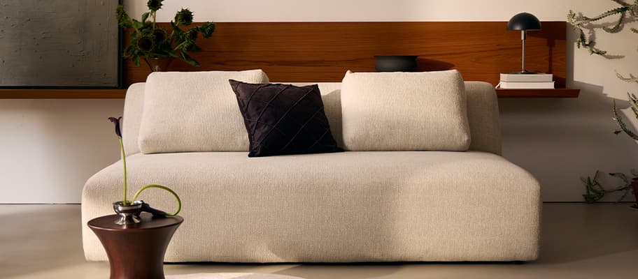 Beiges Sofa mit zwei Rückenpolstern und dunklem Zierkissen vor Holzablage mit Lampe, Büchern und Pflanzen