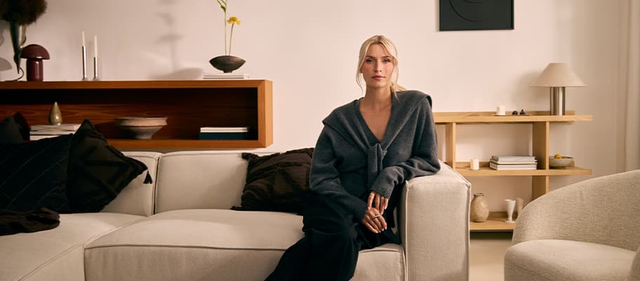 Frau mit grauem Cardigan sitzt auf hellem Sofa in modernem Wohnzimmer mit Regalen und Deko im Hintergrund
