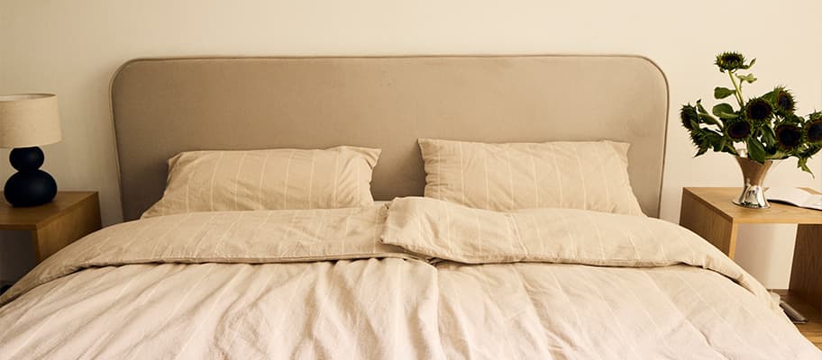 Doppelbett mit beiger Bettdecke und Kissen, Nachttische mit Lampe links und Sonnenblumenstrauß rechts